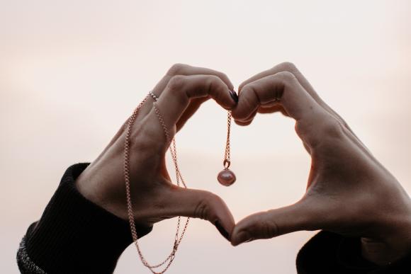 zwei Hände, die ein Herz formen, um eine Hand gewickelte, baumelnde Halskette mit Perlenanhänger, rosa Himmel im Hintergrund